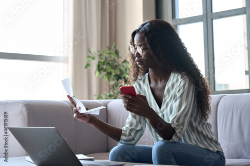 Slika na platnu Serious young black African woman holding paper document calculating rent or money savings, paying bills in mobile application on cell phone doing monthly paperwork sitting on couch at home