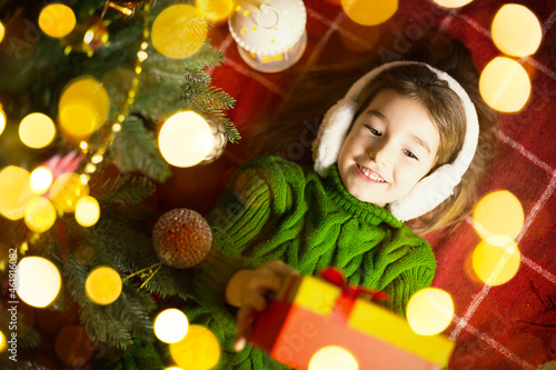 Girl in a warm knitted sweater and fur headphones is lying on a blanket near the Christmas tree with a gift box. Christmas Eve, a dream and a wish. New Year, festive atmosphere, garlands and bokeh