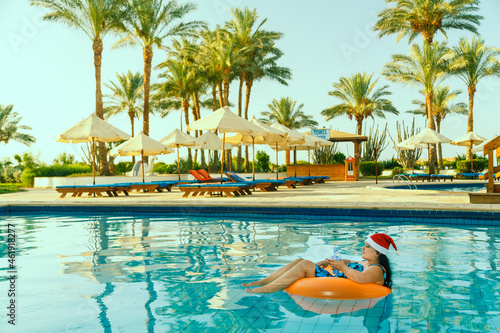 Young brunette woman in santa claus hat in a swimming circle in the pool in the Christmas holidays on the background of palm trees.