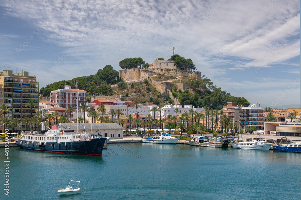 Denia Castle in sunshine with beautiful clouds. In front of it are the houses of the Promeandes. In the foreground is the harbor with ships and fishing boats. A historic ship is moored on the left.