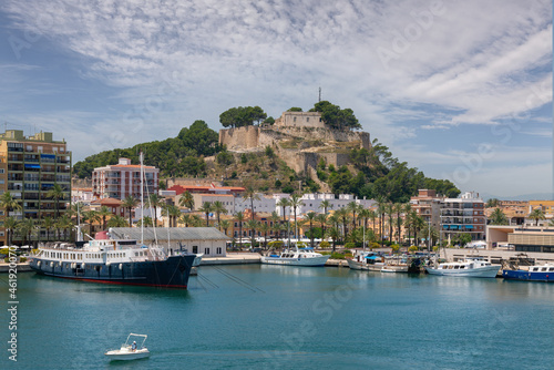 Denia Castle in sunshine with beautiful clouds. In front of it are the houses of the Promeandes. In the foreground is the harbor with ships and fishing boats. A historic ship is moored on the left.