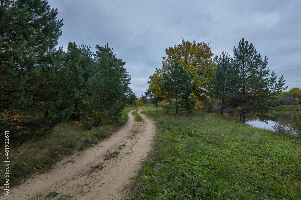 Dirt road along the river bank. Cloudy autumn weather. Rural landscape with road, river and autumn forest.