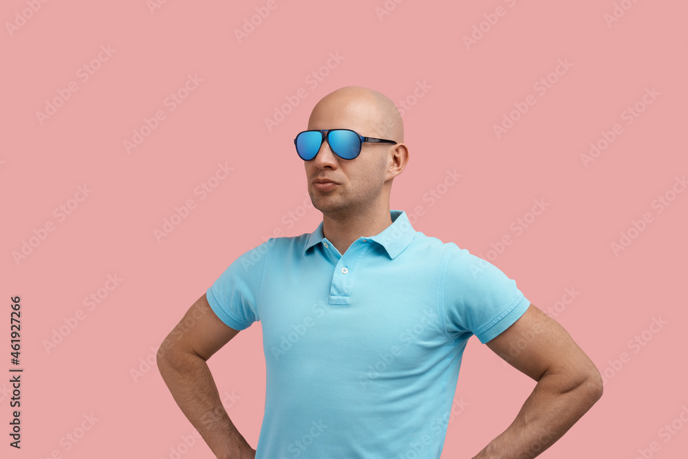 Serious handsome bald homosexual man with bristle looks aside, keeps hands on waist, stays akimbo, needs to decide, gay friendly, wears blue polo shirt and sunglasses, stands on pink background.