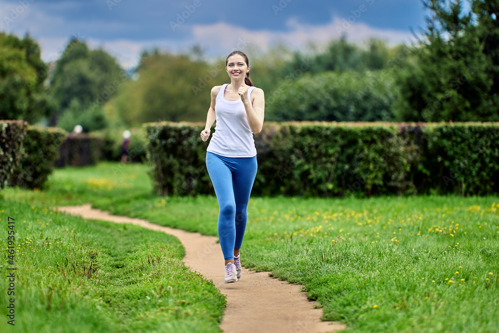 Happy woman in sportswear is running on path in park.