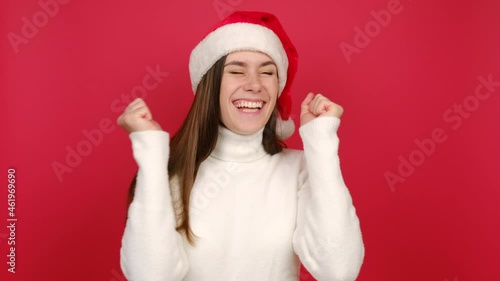 Winner young woman in white cozy sweater and Santa Christmas hat try to hear you overhear listening intently with hand near ear, isolated on red studio background wall. Happy New Year holiday concept photo