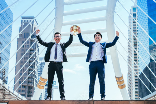 Successful businessman celebrating his victory with arms up and holding gold trophy on cityscape background.
