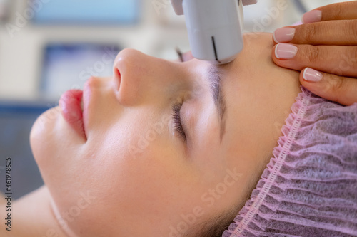 Woman in a spa salon on cosmetic procedures for facial care. Cosmetologist making a woman a therapeutic laser processing on a face. Beautician makes laser procedures on a female face.
