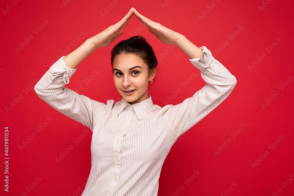 Photo of young positive happy beautiful brunette woman with sincere emotions wearing white shirt isolated over red background with copy space and and making house gesture with hands Photo of young positive happy beautiful brunette woman with sincere emotions wearing white shirt isolated over red background with copy space and and making house gesture with hands