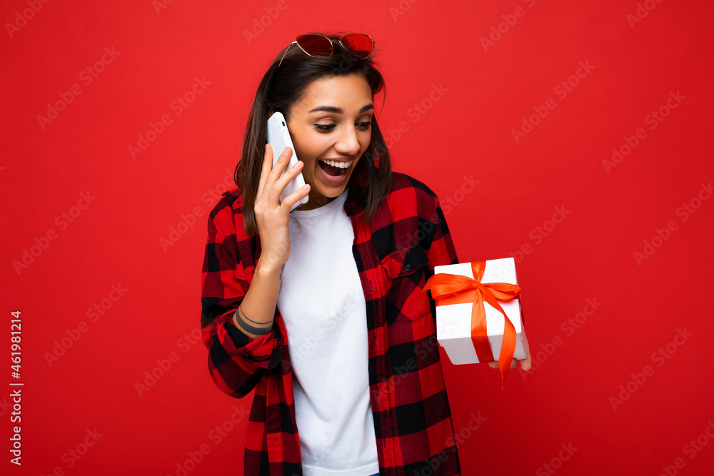 Photo shot of pretty positive surprised young brunet woman isolated over colourful background wall wearing trendy outfit look holding gift box and looking at present box with red ribbon Photo shot of pretty positive surprised young brunet woman isolated over colourful background wall wearing trendy outfit look holding gift box and looking at present box with red ribbon