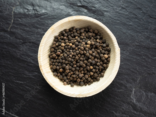 Whole black peppercorns in bamboo bowl on slate worktop, background. Flat lay, top view.
