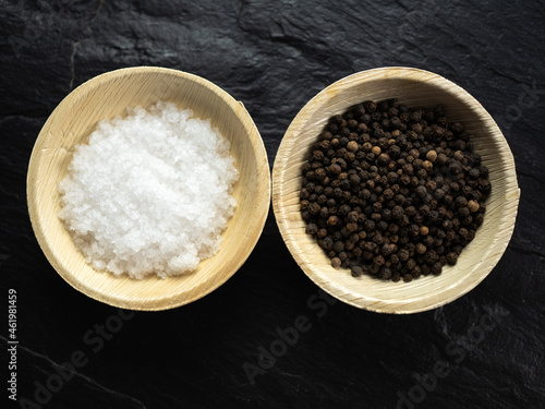 Coarse sea salt and whole black peppercorns in bamboo bowls on slate worktop, background. Flat lay, top view.
