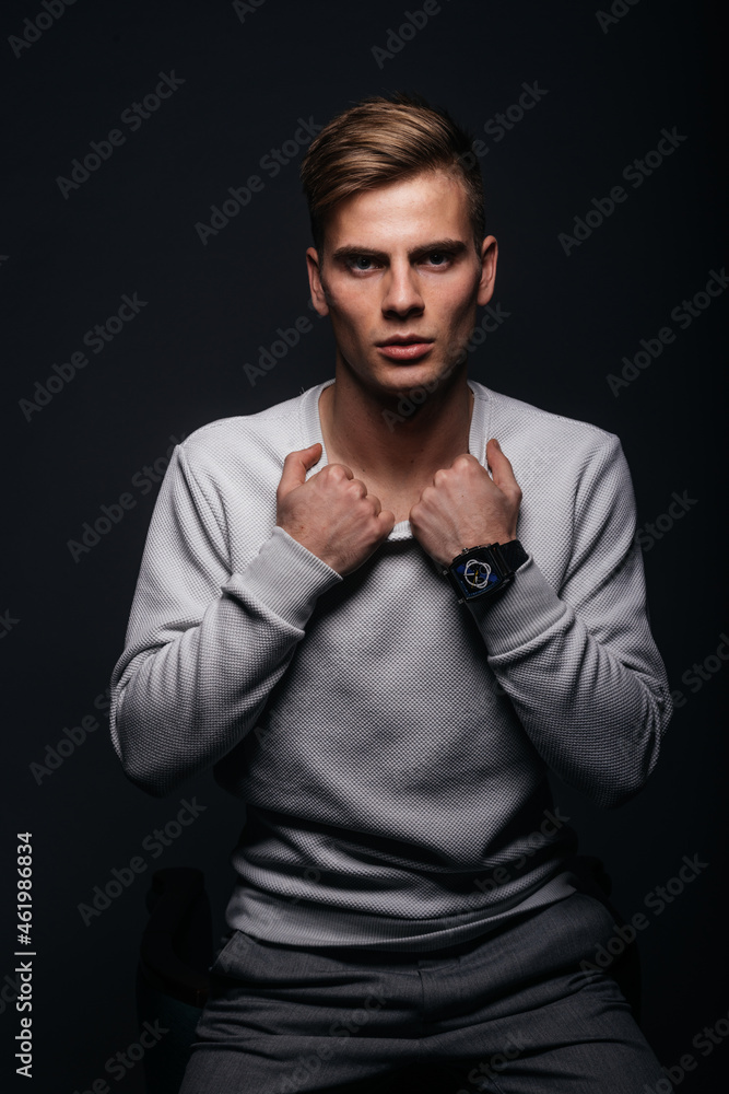 Obraz premium Informally ( casual ) dressed blonde young man with sharp jawline in his 20's posing in a studio in front of a black background while wearing a white sweater.