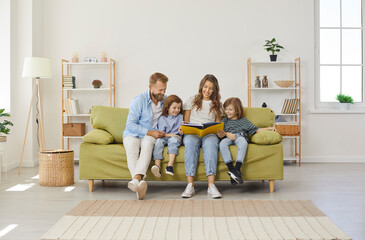 Happy family reading book at home. Mom, dad, little kids sitting together on green sofa in new big light living room with white walls, beige brown rug, wicker storage basket, and shelves in background