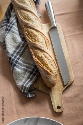 Mediterranean french fresh baguette over a wooden cutting board next to a kitchen napkin and a metallic bread-knife.
