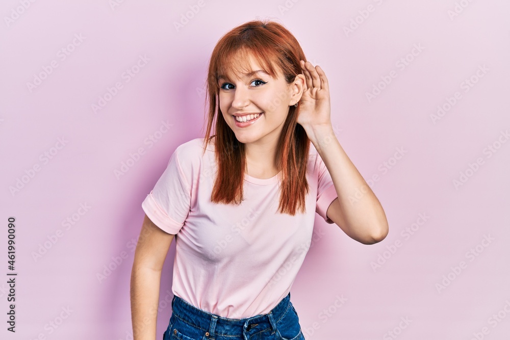 Redhead young woman wearing casual pink t shirt smiling with hand over ear listening an hearing to rumor or gossip. deafness concept. Redhead young woman wearing casual pink t shirt smiling with hand over ear listening an hearing to rumor or gossip. deafness concept.