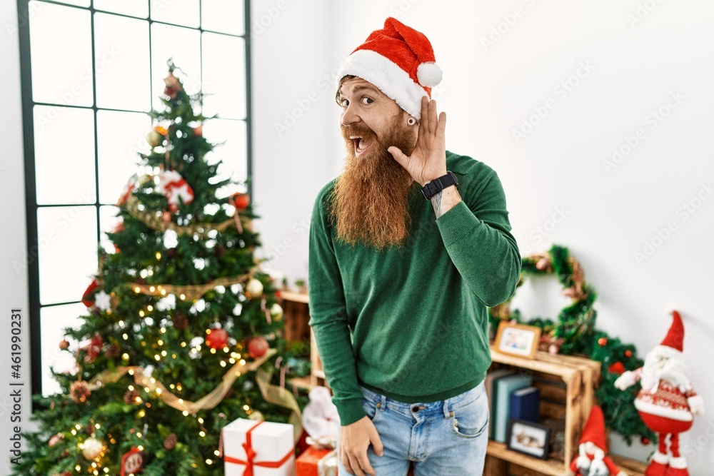 Redhead man with long beard wearing christmas hat by christmas tree smiling with hand over ear listening an hearing to rumor or gossip. deafness concept. Redhead man with long beard wearing christmas hat by christmas tree smiling with hand over ear listening an hearing to rumor or gossip. deafness concept.