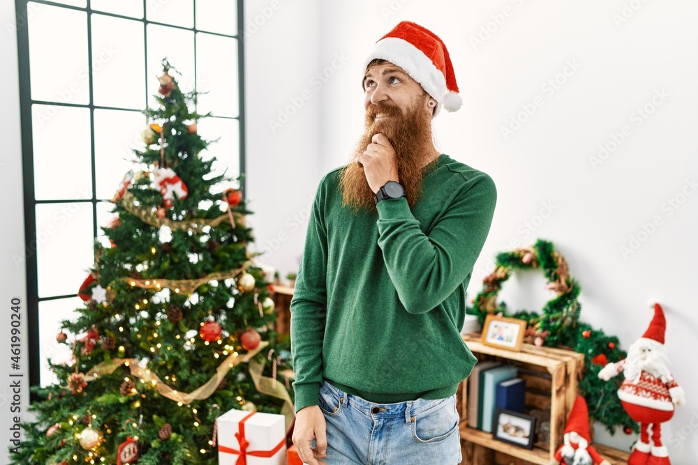 Redhead man with long beard wearing christmas hat by christmas tree thinking worried about a question, concerned and nervous with hand on chin Redhead man with long beard wearing christmas hat by christmas tree thinking worried about a question, concerned and nervous with hand on chin