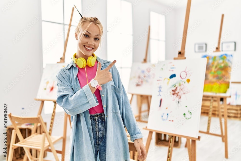 Naklejka premium Young caucasian girl at art studio cheerful with a smile on face pointing with hand and finger up to the side with happy and natural expression