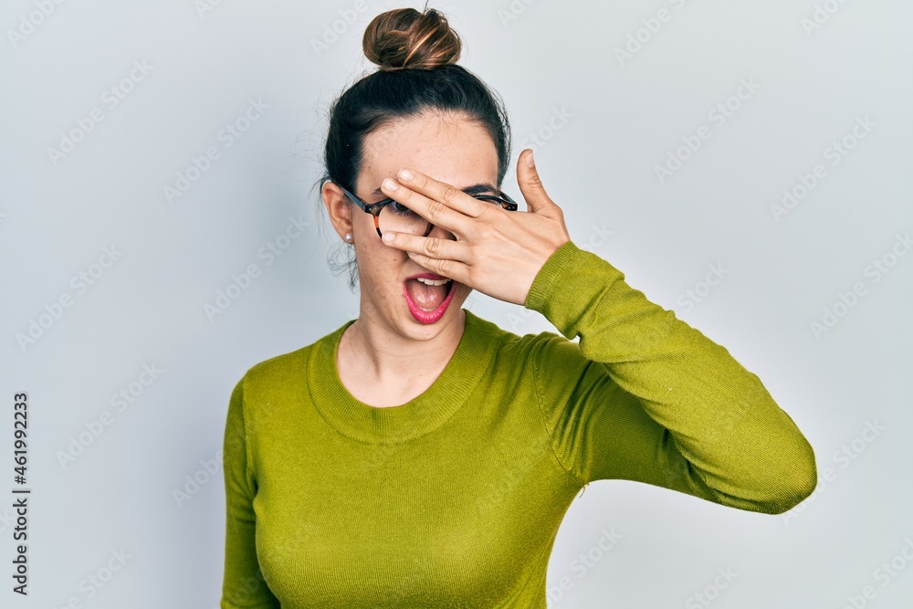 Young hispanic girl wearing casual clothes and glasses peeking in shock covering face and eyes with hand, looking through fingers with embarrassed expression. Young hispanic girl wearing casual clothes and glasses peeking in shock covering face and eyes with hand, looking through fingers with embarrassed expression.