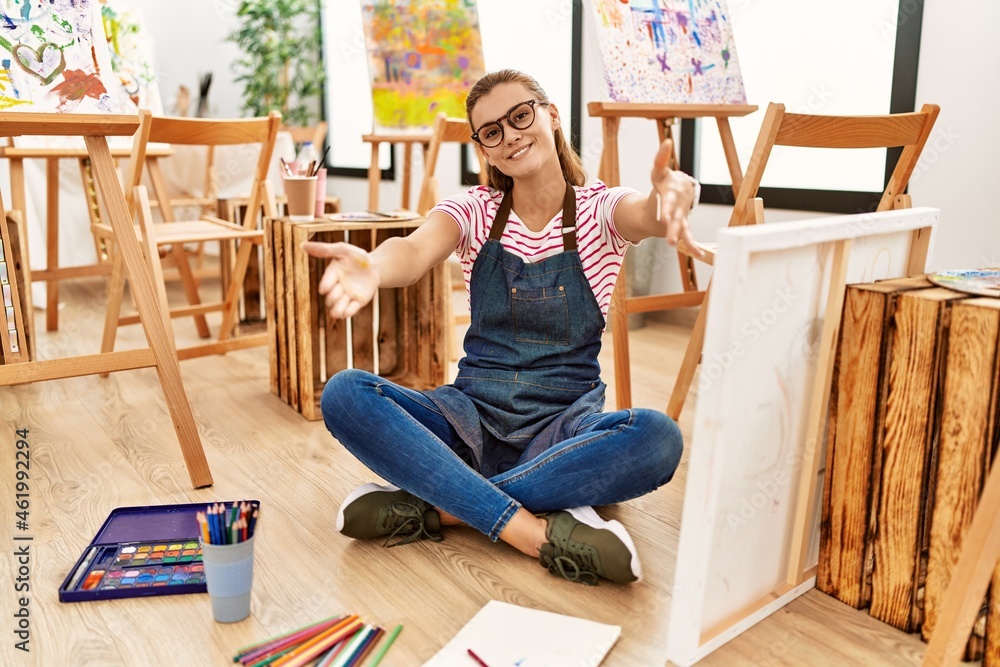 Young brunette woman at art studio sitting on the floor looking at the camera smiling with open arms for hug. cheerful expression embracing happiness.