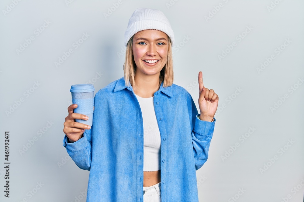 Beautiful blonde woman drinking a take away cup of coffee smiling with an idea or question pointing finger with happy face, number one Beautiful blonde woman drinking a take away cup of coffee smiling with an idea or question pointing finger with happy face, number one
