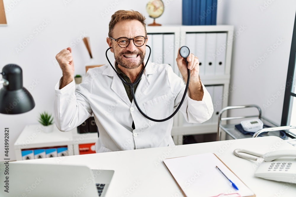 Middle age man with beard wearing doctor uniform holding stethoscope screaming proud, celebrating victory and success very excited with raised arm