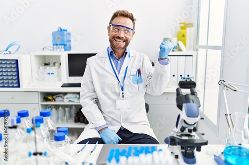 Middle age man working at scientist laboratory smiling with happy face looking and pointing to the side with thumb up.