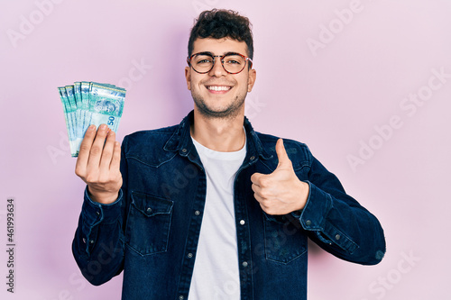 Young hispanic man holding 50 malaysia ringgit banknotes smiling happy and positive, thumb up doing excellent and approval sign photo