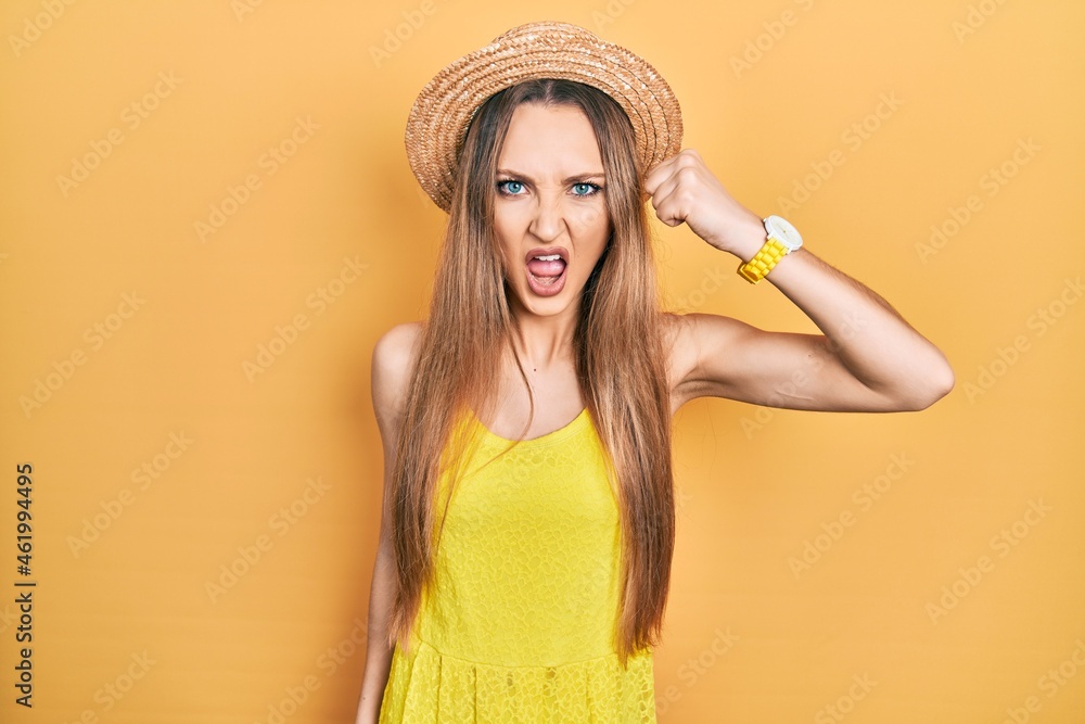 Young blonde girl wearing summer hat angry and mad raising fist frustrated and furious while shouting with anger. rage and aggressive concept. Young blonde girl wearing summer hat angry and mad raising fist frustrated and furious while shouting with anger. rage and aggressive concept.