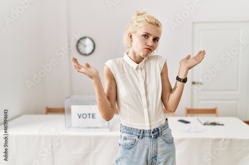 Photos Beautiful caucasian woman standing by voting ballot at election room clueless and confused expression with arms and hands raised