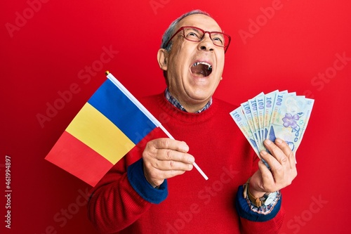 Handsome senior man with grey hair holding romania flag and leu banknotes angry and mad screaming frustrated and furious, shouting with anger looking up.