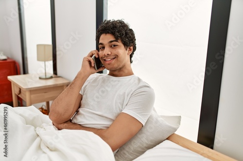 Young hispanic man talking on the smartphone sitting on the bed at bedroom.