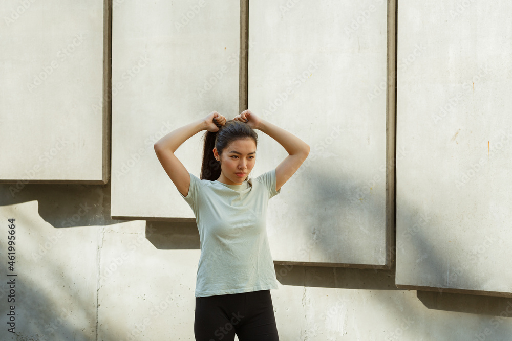 Fototapeta premium Pretty Asian young woman in t-shirt adjusts ponytail made of long dark hair standing near white wall on sunny day middle view