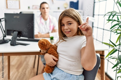 Blonde little girl at pediatrician clinic with female doctor surprised with an idea or question pointing finger with happy face, number one