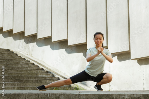 Attractive Asian lady in sportswear and sneakers does side dynamic lunges near underground crossing steps and street wall