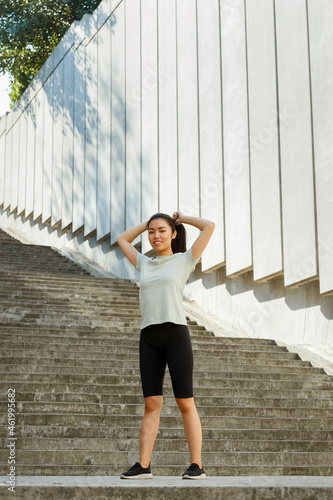 Pretty Asian young woman in t-shirt adjusts ponytail made of long dark hair standing near white wall on sunny day close view