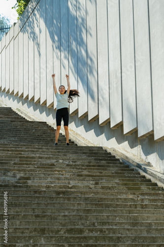 Joyful Asian lady in tracksuit raises up hands standing on underground crossing stone stairs by wall with tree shadow in summer © sergiymolchenko