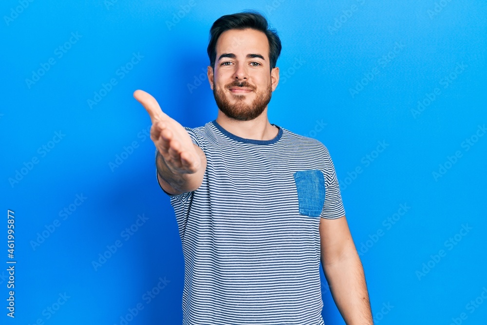 Handsome caucasian man with beard wearing casual striped t shirt smiling friendly offering handshake as greeting and welcoming. successful business. Handsome caucasian man with beard wearing casual striped t shirt smiling friendly offering handshake as greeting and welcoming. successful business.