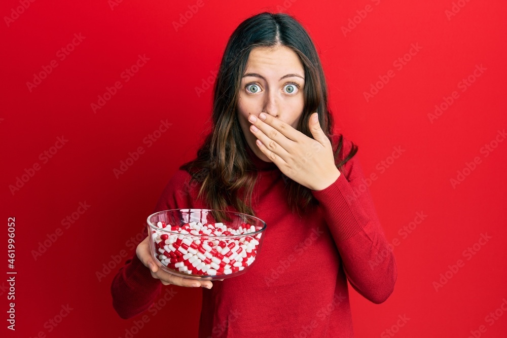 Young brunette woman holding bowl full of pills covering mouth with hand, shocked and afraid for mistake. surprised expression Young brunette woman holding bowl full of pills covering mouth with hand, shocked and afraid for mistake. surprised expression