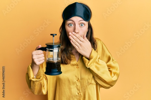 Photos Young brunette woman wearing sleep mask holding coffee maker covering mouth with hand, shocked and afraid for mistake