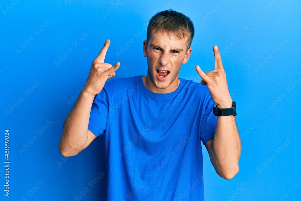 Young caucasian man wearing casual blue t shirt shouting with crazy expression doing rock symbol with hands up. music star. heavy music concept. Young caucasian man wearing casual blue t shirt shouting with crazy expression doing rock symbol with hands up. music star. heavy music concept.