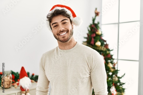 Young hispanic man smiling happy wearing christmas hat standing at home.