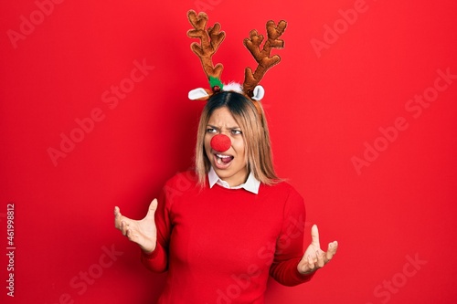 Beautiful hispanic woman wearing deer christmas hat and red nose crazy and mad shouting and yelling with aggressive expression and arms raised. frustration concept.