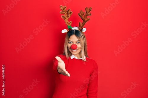 Beautiful hispanic woman wearing deer christmas hat and red nose smiling friendly offering handshake as greeting and welcoming. successful business.