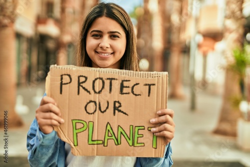 Young latin girl smiling happy holding protect our planet banner at the city.