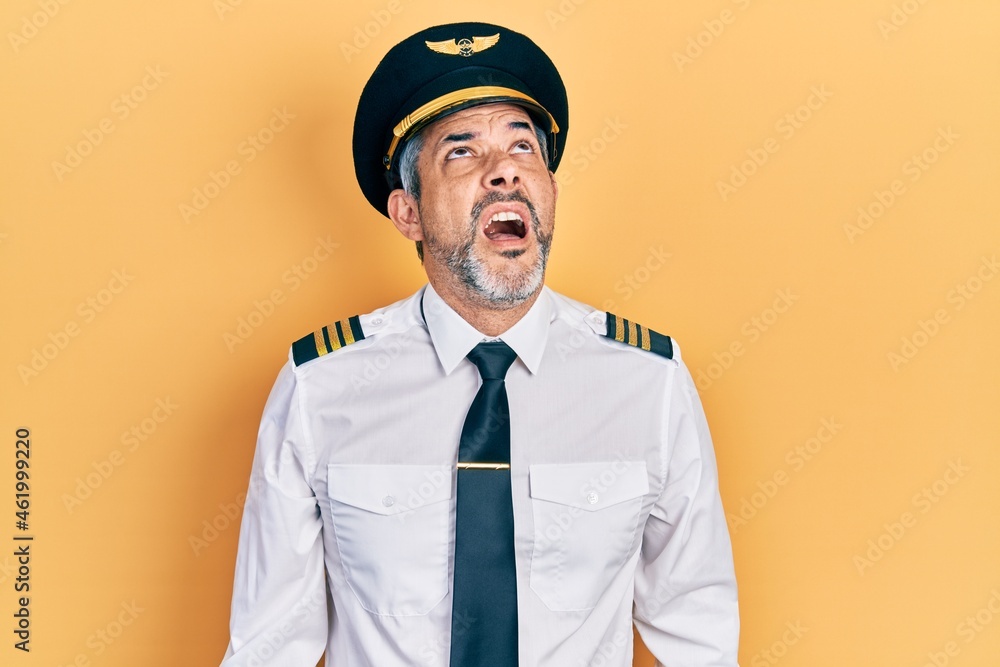 Handsome middle age man with grey hair wearing airplane pilot uniform angry and mad screaming frustrated and furious, shouting with anger. rage and aggressive concept. Handsome middle age man with grey hair wearing airplane pilot uniform angry and mad screaming frustrated and furious, shouting with anger. rage and aggressive concept.