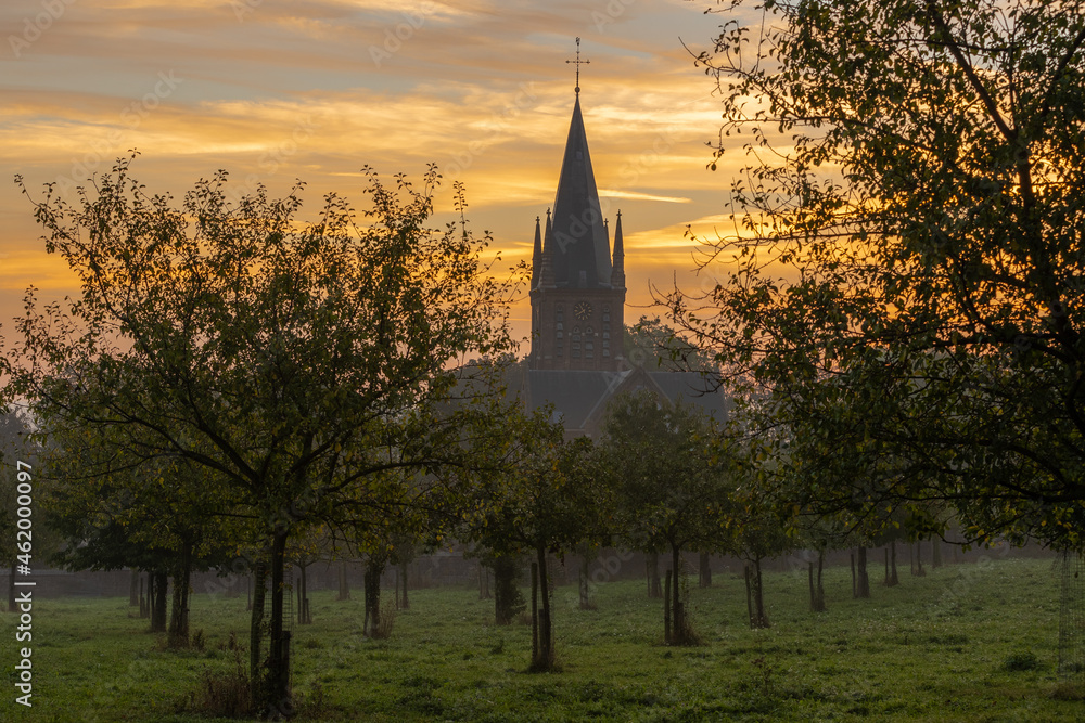 Obraz premium A typical colorful Autumn sunrise in Maastricht with the landscape covered with a layer of fog, leaving only silhouettes visible in the distance, like the church on the hillside accompanied with trees
