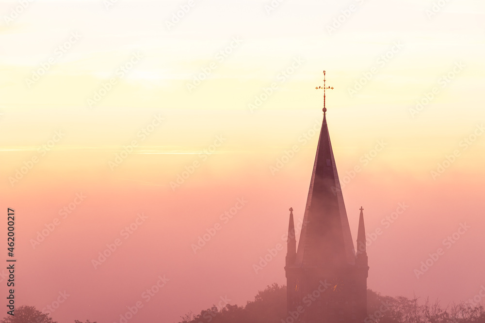 Fototapeta premium A typical colorful Autumn sunrise in Maastricht with the landscape covered with a layer of fog, leaving only silhouettes visible in the distance, like this tower of a church on the hillside.