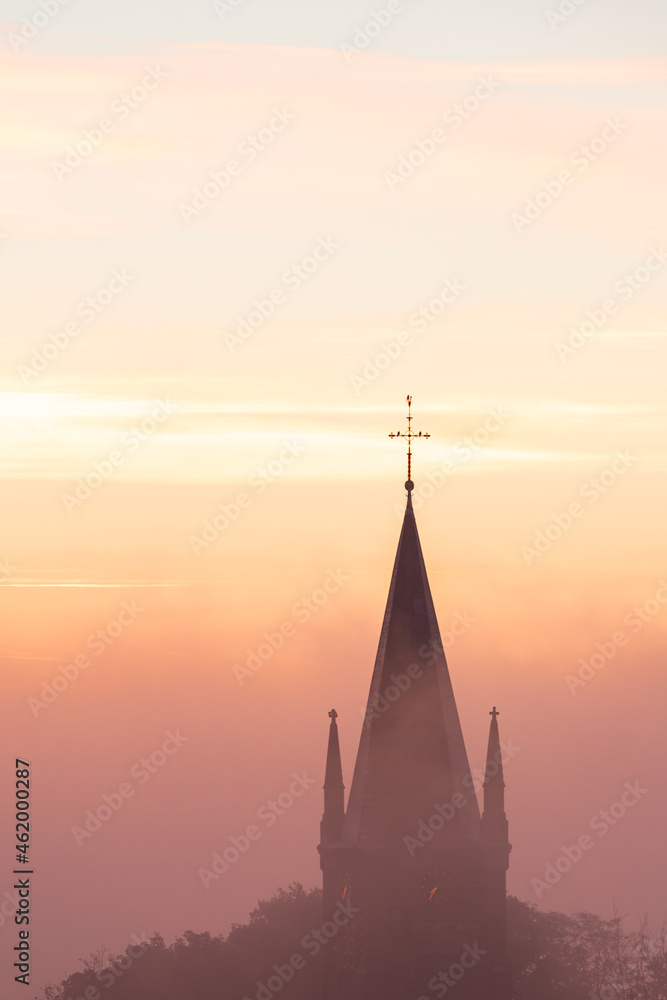 Fototapeta premium A typical colorful Autumn sunrise in Maastricht with the landscape covered with a layer of fog, leaving only silhouettes visible in the distance, like this tower of a church on the hillside.