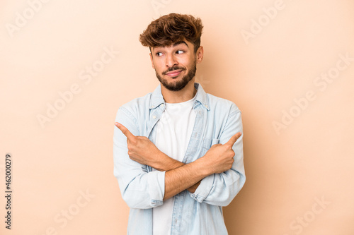 Young arab man isolated on beige background points sideways, is trying to choose between two options. © Asier